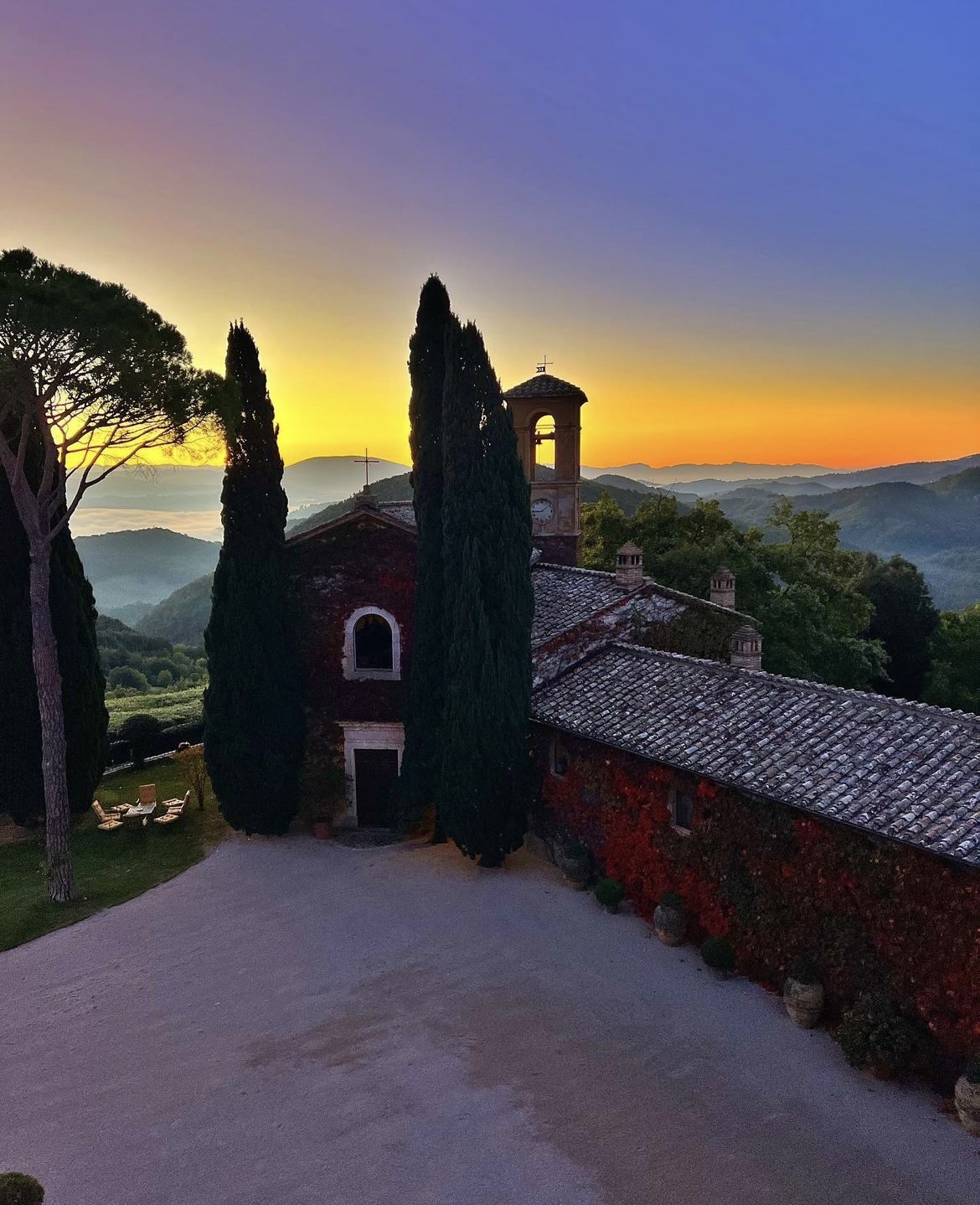 A stone villa and its bell tower among cypresses, the walls covered in red autumn ivy, with mist in the valleys at sunrise