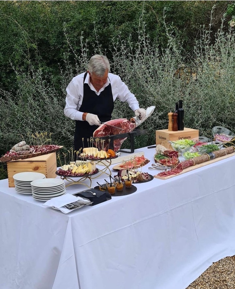 A chef in a black apron carving prosciutto at an outdoor table laid with cheeses, salumi and salads in a villa garden