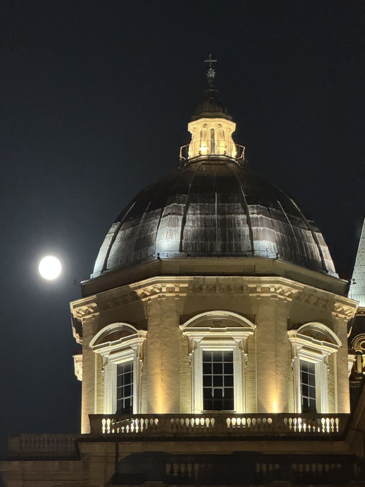 A church dome lit up at night with the full moon beside it