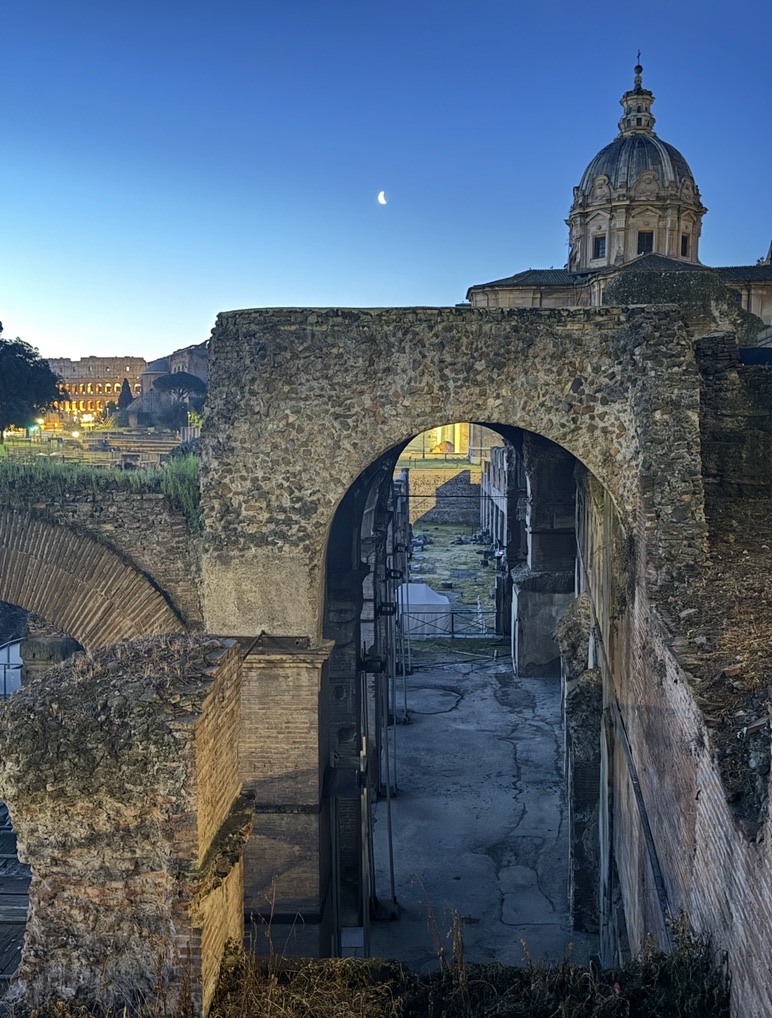 A stone arch in the Roman Forum at dusk, with a crescent moon, a church dome and the Colosseum lit in the distance