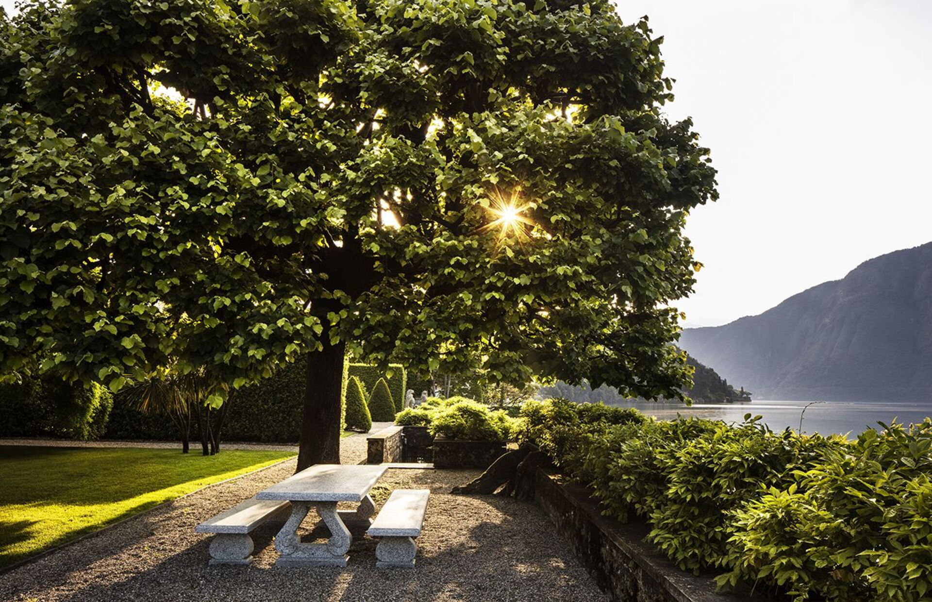 Stone garden bench under a sun-dappled plane tree at Villa Balbiano with Lake Como and the western shore in the background