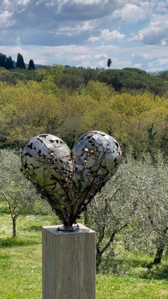 A steel heart sculpture on a stone plinth in an olive grove, with Tuscan hills and cypresses behind