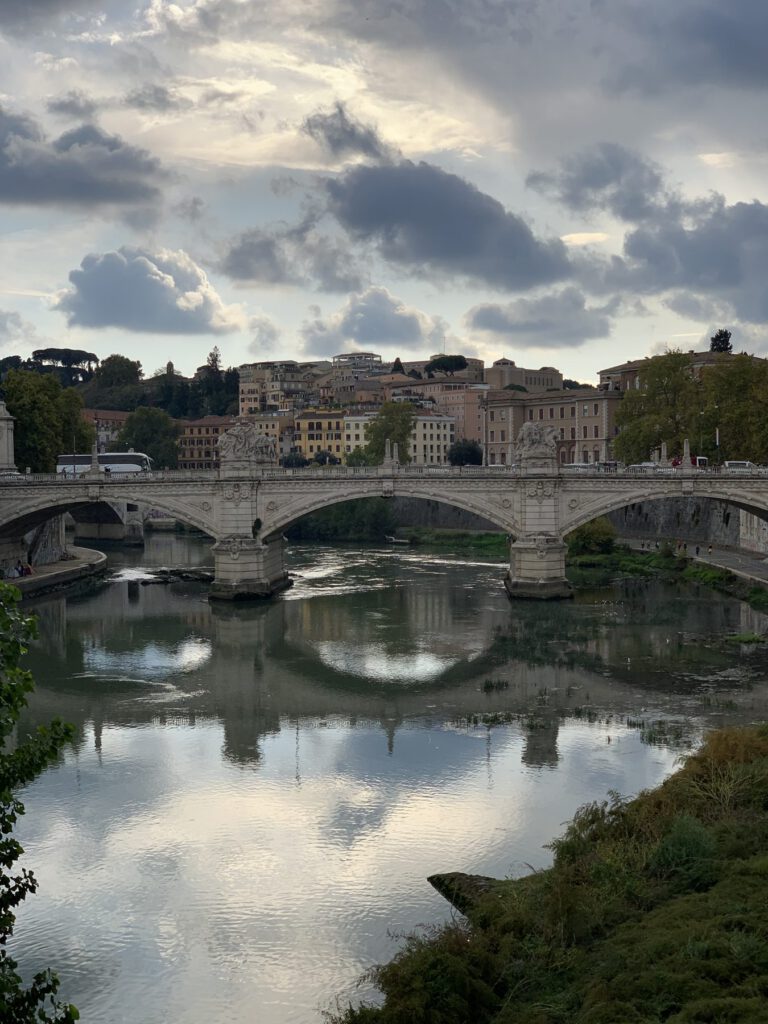 A stone bridge over the Tiber in Rome reflected in the still river under a cloudy sky