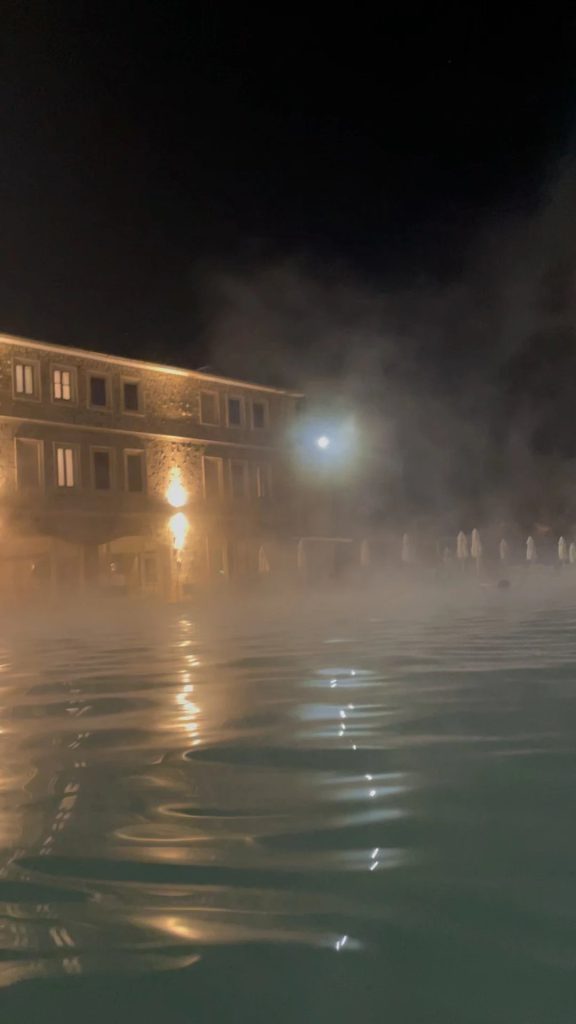 Steam rising from the thermal pool at Saturnia on a cool night, with the lit stone building behind