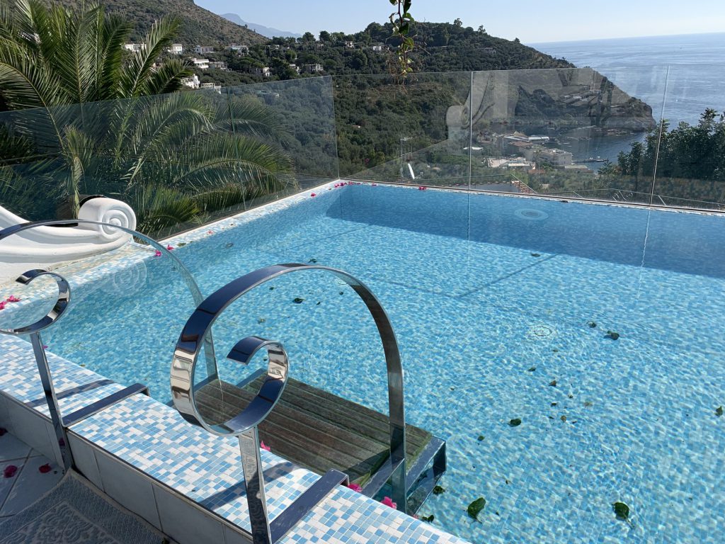 An infinity pool on a villa terrace with a glass railing, looking out over the hills and the sea